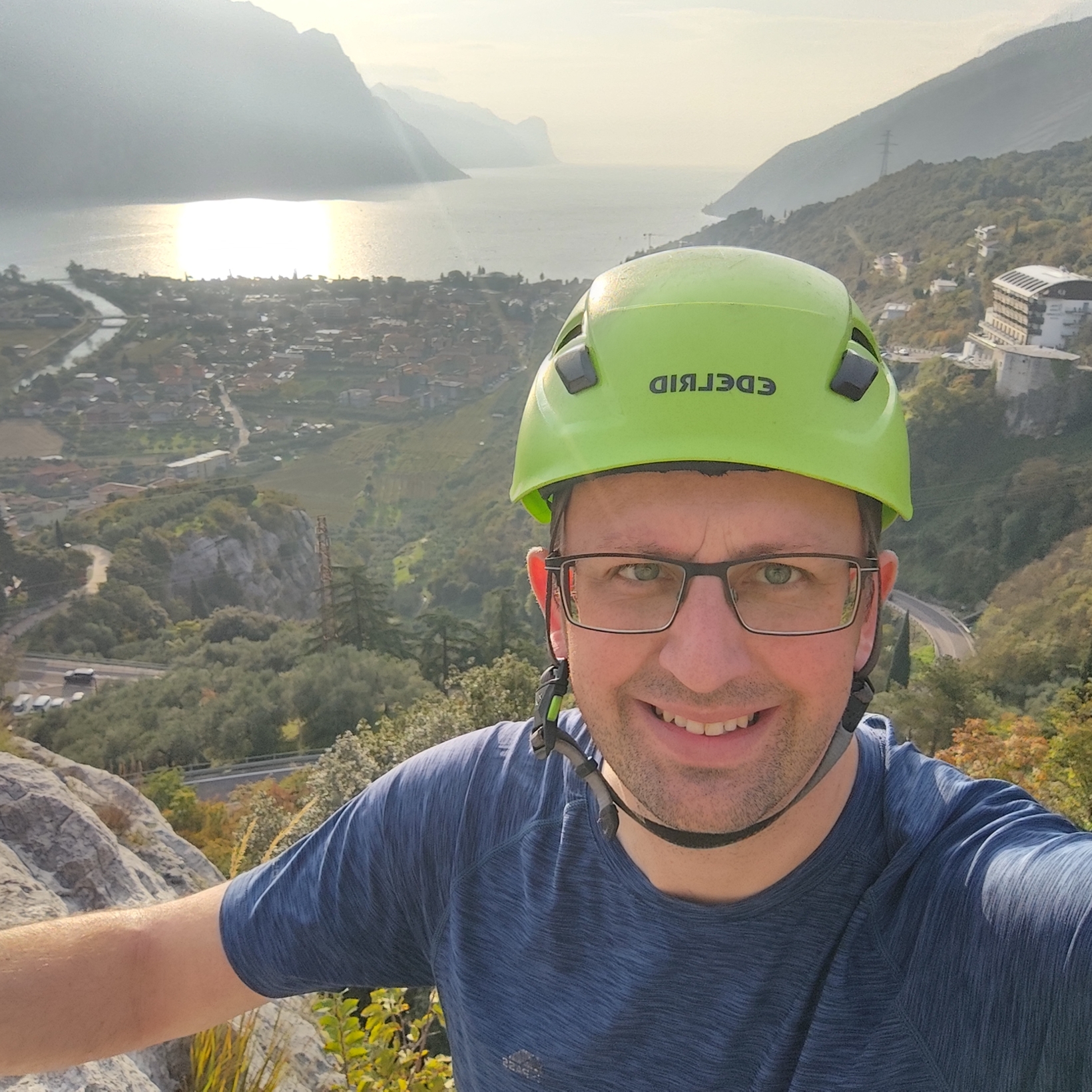 Auf dem Bild ist ein Mann mit einem grünen Helm zu sehen, der sich in einer bergigen Landschaft mit Blick auf einen See und eine Stadt befindet.