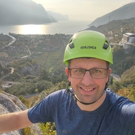 Auf dem Bild ist ein Mann mit einem grünen Helm zu sehen, der sich in einer bergigen Landschaft mit Blick auf einen See und eine Stadt befindet.